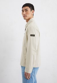Beige lightweight jacket with a straight cut, featuring a collar, front pockets, and a dark label on the sleeve. Worn with blue jeans.