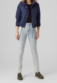 Mörkblå jacka med elastiska ärmar och dragsko, ihop med ljusblå åtsittande jeans och gröna loafers, mot en neutral bakgrund.