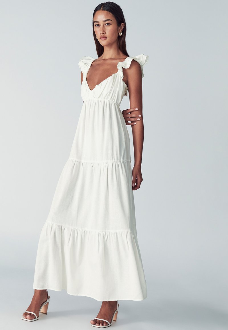 Nainen, joka pukeutuu olkaimettomaan valkoiseen maksimekkoon, jossa on frillaolkaimet ja kerroksellinen helma, yhdistettynä valkoisiin korkokenkiin, seisoo tasaisen taustan edessä.