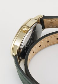 Montre ronde dorée avec un bracelet en cuir noir, présentant une surface texturée et un dos en argent gravé. Dessous vert.