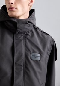 Jachetă impermeabilă de culoare gri închis, cu guler înalt, accente cu nasturi și o porțiune texturată cu logo. Material neted cu un design elegant.