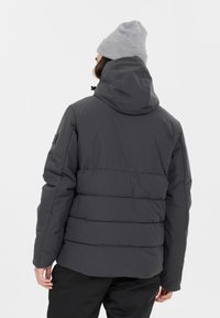 Dunkelgraue Pufferjacke mit Kapuze, mit gesteppten Abschnitten und einem schwarzen Patch am Ärmel. Getragen mit einer hellgrauen Mütze.