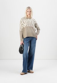 Beige sweater with white zebra stripes, paired with blue wide-leg jeans. Holding a small black handbag. Soft texture, relaxed fit.