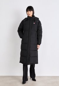 Cappotto puffo nero con design lungo fino al ginocchio, texture trapuntata e chiusura con zip. Presenta un cappuccio e un dettaglio del logo sul petto.