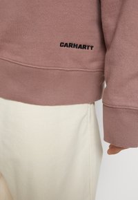 Sudadera rosa clara con puños y dobladillo acanalados, con un logo negro de "CARHARTT" bordado en el lado inferior, combinada con una falda color crema.