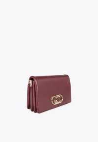 Burgundská kožená kabelka na zápěstí se zlatým emblémem, sklopená s elegantním designem, vystavená na neutrálním pozadí.
