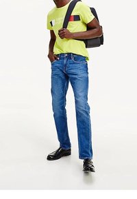 Jeans de mezclilla azul con un corte recto, combinados con una camisa de manga corta de color verde neón que presenta rayas de colores y un logo, y zapatos negros.