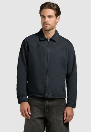 Mężczyzna z jasnym zarostem, ubrany w czarną kurtkę na zamek i ciemnoszare jeansy, stojący na tle jednolitego jasnego tła.