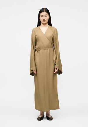 Femme aux longs cheveux noirs portant une robe portefeuille beige longue avec des manches larges et des chaussures plates noires, debout devant un fond uni.
