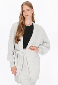 Lichtgrijze gebreide cardigan met een zelfbindende riem, lange mouwen en een zachte textuur, gelaagd over een zwarte top.