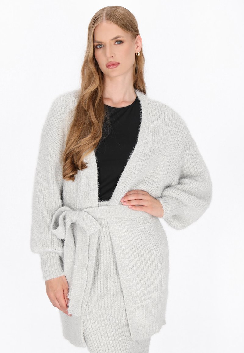 Lichtgrijze gebreide cardigan met een zelfbindende riem, lange mouwen en een zachte textuur, gelaagd over een zwarte top.