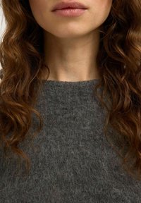 Mujer con cabello rizado castaño que lleva un suéter gris oscuro y peludo, mostrando los labios, la barbilla, el cuello y la parte superior del pecho.