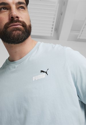 Camiseta de algodón azul claro con mangas cortas, que presenta un logo negro de Puma y texto blanco en el pecho. Textura suave, ajuste casual.