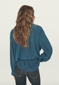 Langærmet, teal bluse lavet af blødt stof, med en afslappet pasform og indsamlede detaljer ved skuldrene, kombineret med højtaljede mørke jeans.