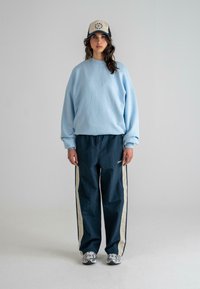 Helles blaues Oversize-Sweatshirt, dunkelblaue Jogginghose mit seitlichen Streifen, silberne Laufschuhe und eine beige Kappe mit navy Akzenten.