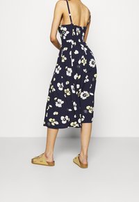 Marineblaues Blumenkleid in Midi-Länge mit verstellbaren Trägern und figurbetontem Oberteil. Es hat ein Muster aus weißen und gelben Blumen und einen ausgestellten Rock. Trägt dazu beige Sandalen.