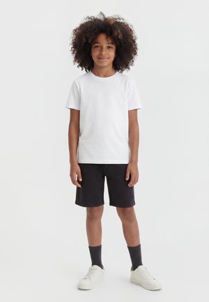 Barn med krøllet hår står vendt fremad, iført en hvid T-shirt, sorte knælange shorts, mørke sokker og hvide sneakers på hvid baggrund.