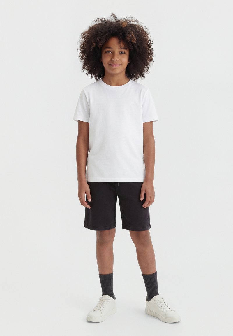 Enfant aux cheveux bouclés, debout face à nous, portant un T-shirt blanc, un short noir jusqu'aux genoux, des chaussettes foncées et des baskets blanches sur fond blanc.