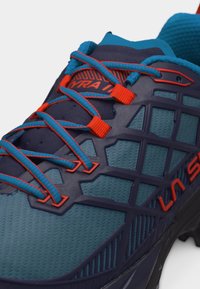 Κοντινό πλάνο σε μπλε και πορτοκαλί παπούτσι για τρέξιμο σε μονοπάτια με λεπτομερή κορδόνια, πλέγμα στο πάνω μέρος και ενισχυμένα τμήματα κοντά στα δάχτυλα και στα πλαϊνά.