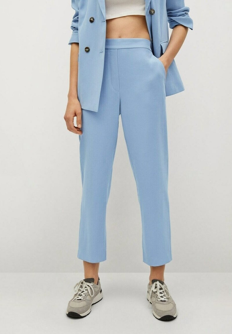 Personne portant un pantalon court bleu clair et un blazer assorti avec un crop top blanc et des baskets beige sur un fond uni.