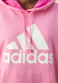 Rosa hoodie i mjukt material med en stor vit Adidas-logotyp över bröstet. Inkluderar huva med dragsko och en framficka.