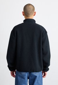 Czarna bluza polarowa z wysokim kołnierzem, o fakturowanej powierzchni, luźnym kroju i długich rękawach, zestawiona z niebieskimi dżinsami, widziana od tyłu.