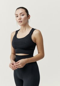 Musta urheiluliivi, jossa on syvä pääntie, tekstuuriset yksityiskohdat ja leveät hihnat, yhdistettynä korkeavyötäröisiin mustiin legginsseihin, joissa on sileä pinta.