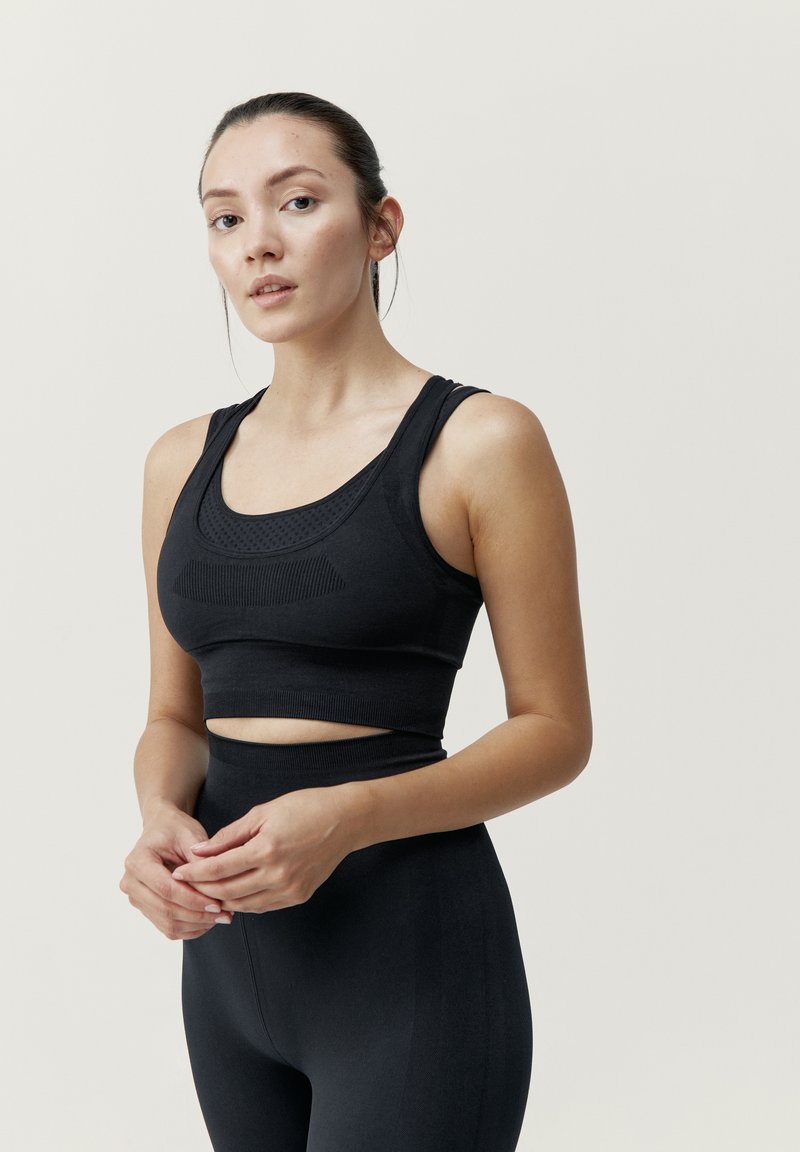 Musta urheiluliivi, jossa on syvä pääntie, tekstuuriset yksityiskohdat ja leveät hihnat, yhdistettynä korkeavyötäröisiin mustiin legginsseihin, joissa on sileä pinta.