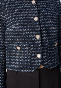 Gros plan d'une veste en tweed bleu et noir avec un tissu texturé et des boutons décoratifs dorés sur un pantalon noir.