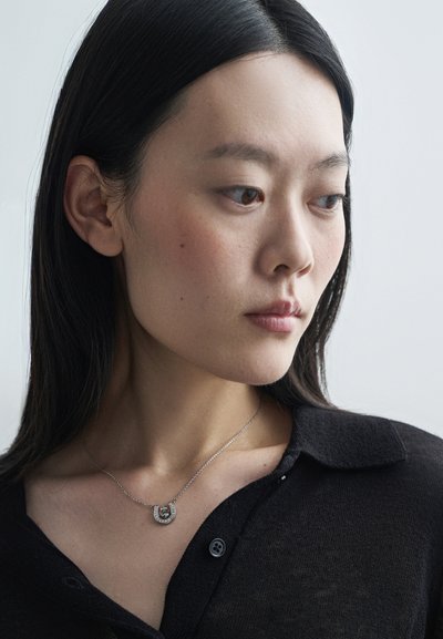 Collar de plata con un colgante en forma de herradura, que presenta pequeñas piedras incrustadas, llevado sobre una blusa de punto negra con cuello. Textura suave y ligero brillo.