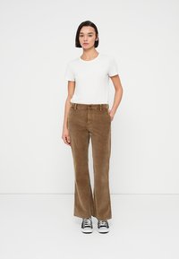 Καφέ παντελόνι καμπάνα από κορντόνι με μπροστινές τσέπες, σε συνδυασμό με ένα απλό λευκό t-shirt και μαύρα αθλητικά παπούτσια, στέκεται μπροστά σε απλό φόντο.