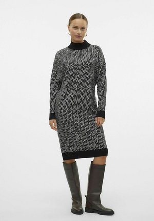 Robe en tricot noir et blanc avec un motif géométrique, manches longues et col montant. Portée avec des bottes noires montantes.