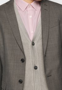 Giacca da suit in misto lana grigia con revers a notch, abbinata a un cardigan grigio chiaro e a una camicia button-up rosa pallido. I bottoni neri decorano il cardigan.