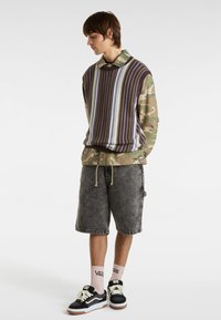 Gestreifter lila- und cremefarbener Pullover über einem camo-farbenen Hemd, kombiniert mit grauen Jeansshorts, rosa Socken und schwarzen Sneakers mit weißen Akzenten.