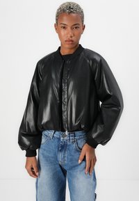 Giubbotto bomber in finta pelle nera con polsini e colletto a coste, con zip frontale e vestibilità rilassata, abbinato a jeans blu a vita alta.