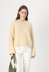 Crème-kleurige trui met een gestructuurd breiwerk, ronde halslijn en brede mouwen, gecombineerd met witte jeans en een groene handtas.