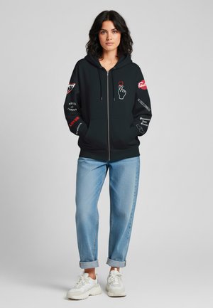 Felpa nera con zip e stampe grafiche in rosso e bianco, con un'icona di mano, frasi e dettagli sulle maniche. Abbinata a jeans con risvolto.