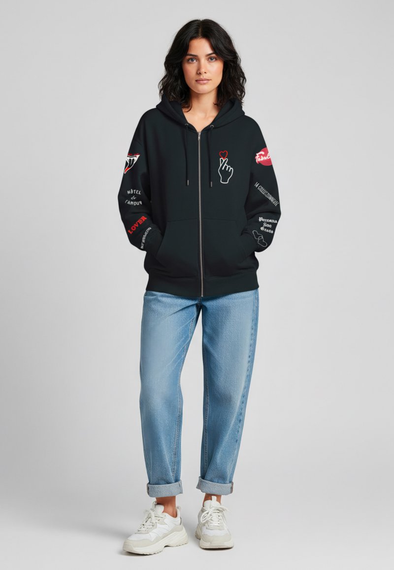 Felpa nera con zip e stampe grafiche in rosso e bianco, con un'icona di mano, frasi e dettagli sulle maniche. Abbinata a jeans con risvolto.