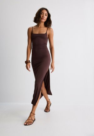 Femme portant une robe marron sans manches à bretelles fines et fente sur le côté, sandales marron et bracelets, debout devant un fond uni.