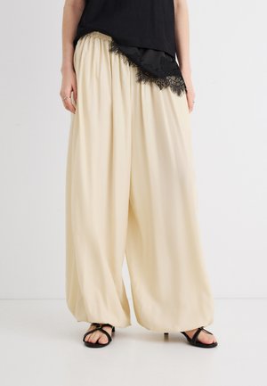 Kvinde iført beige bukser med vide ben og samlet talje, sort asymmetrisk top med blondekanter og sorte sandaler med stropper.