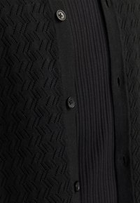 Cardigan nero con motivo a zigzag testurizzato sul davanti, dotato di un pannello a coste e chiusura con bottone nero. Realizzato in morbido tessuto a maglia.