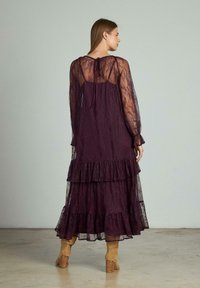 Abito lungo a strati in pizzo viola scuro con maniche sheer lunghe e un nodo nella parte posteriore del collo. Presenta un orlo arruffato e una silhouette fluente.