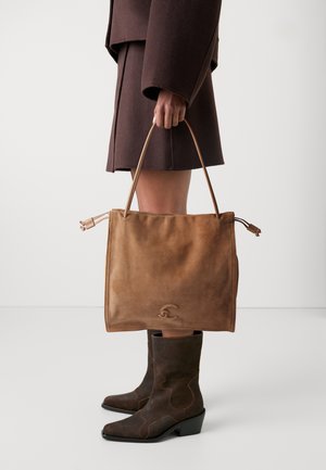 Borsa tote in suede marrone con una texture liscia, caratterizzata da due manici e un logo sottile impresso sul fronte, abbinata a stivali marroni scuri.