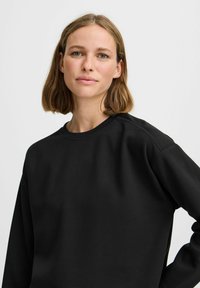 Melna sudžiga, kas izgatavota no kokvilnas maisījuma, ar apaļu kakla izgriezumu, brīvu piegriezumu un iestrādātām piedurknēm. Gluda, mīksta tekstūra bez redzamām rakstām.