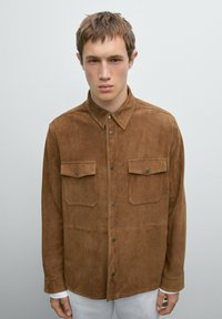 Braunes Wildleder-Hemd mit Knopfleiste hat einen Kragen, zwei Brusttaschen mit Klappen und einen geraden Schnitt. Sanfte Textur, minimalistisches Design.
