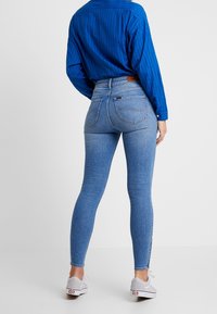 Jeans de mezclilla azules ajustados con una cintura alta, un ligero estiramiento y bolsillos traseros, combinados con una camisa de manga larga a rayas azules y zapatos grises.