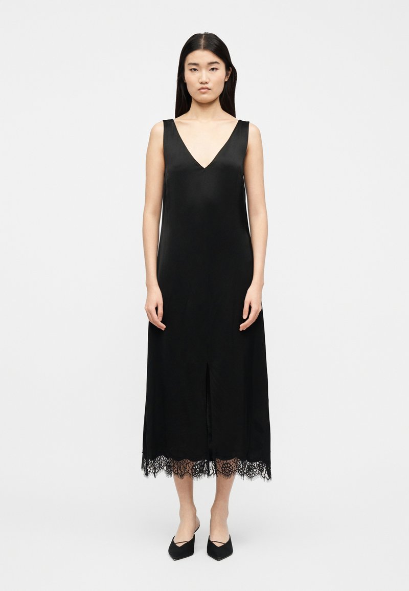 Γυναίκα που φοράει αμάνικο μαύρο μίντι φόρεμα με V λαιμόκοψη και τελείωμα με δαντέλα, όρθια με μαύρες μυτερές μπαλαρίνες σε λευκό φόντο.