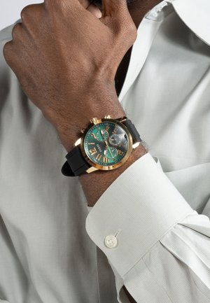 Ruka s zlatými a čiernymi hodinkami so zeleným ciferníkom, oblečená v bielej košeli s gombíkmi, dotýkajúc sa brady.