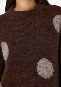 Närbild på en brun ulltröja med stora, fluffiga ljusrosa prickar och en ribbad rund halsringning som bärs av en person.