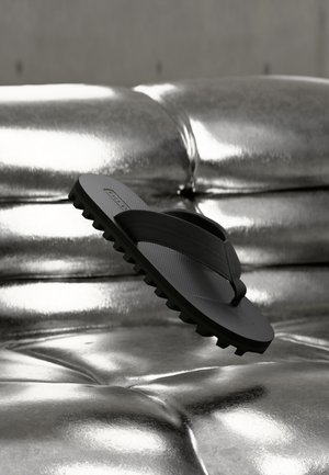 Infradito nero in gomma con suola strutturata che fluttua sopra una superficie argentata metallica lucida e trapuntata, evidenziando il contrasto tra texture e colore.